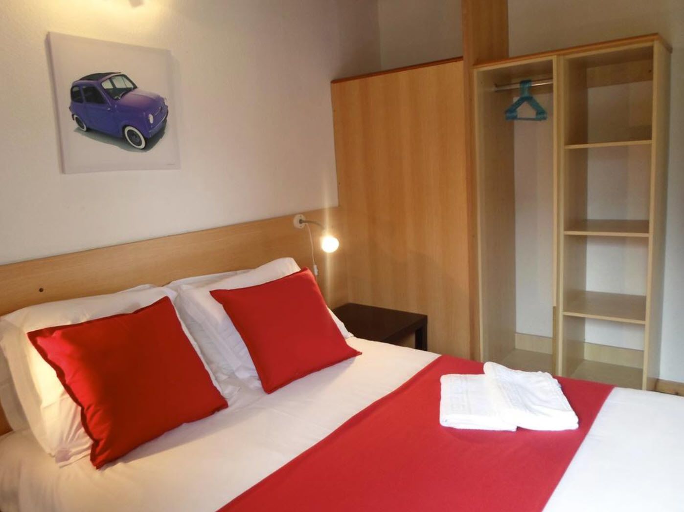 Una habitación de hotel con cama, ropa de cama roja y blanca, cabecera de madera, mesita de noche, lámpara, armario y un cuadro en la pared con una imagen de un coche.