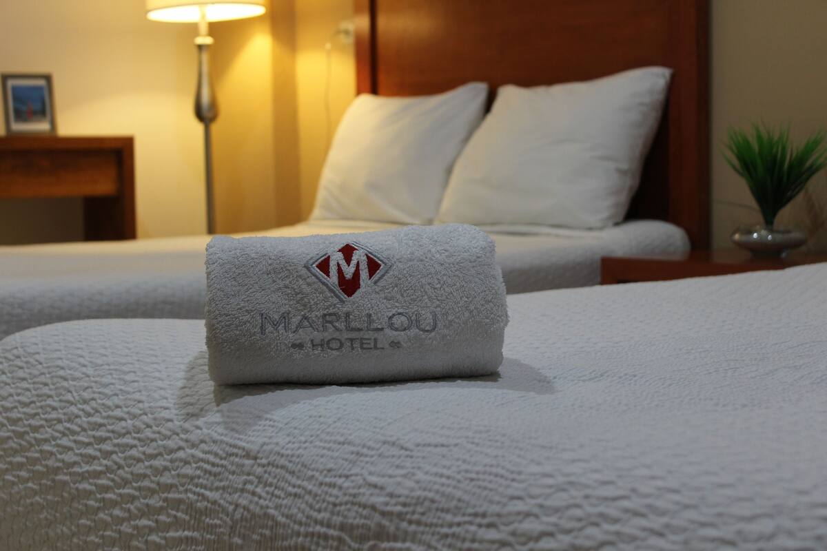 Ein ordentlich bezogenes Bett mit einem gefalteten Handtuch mit dem Logo des Marllou Hotels.