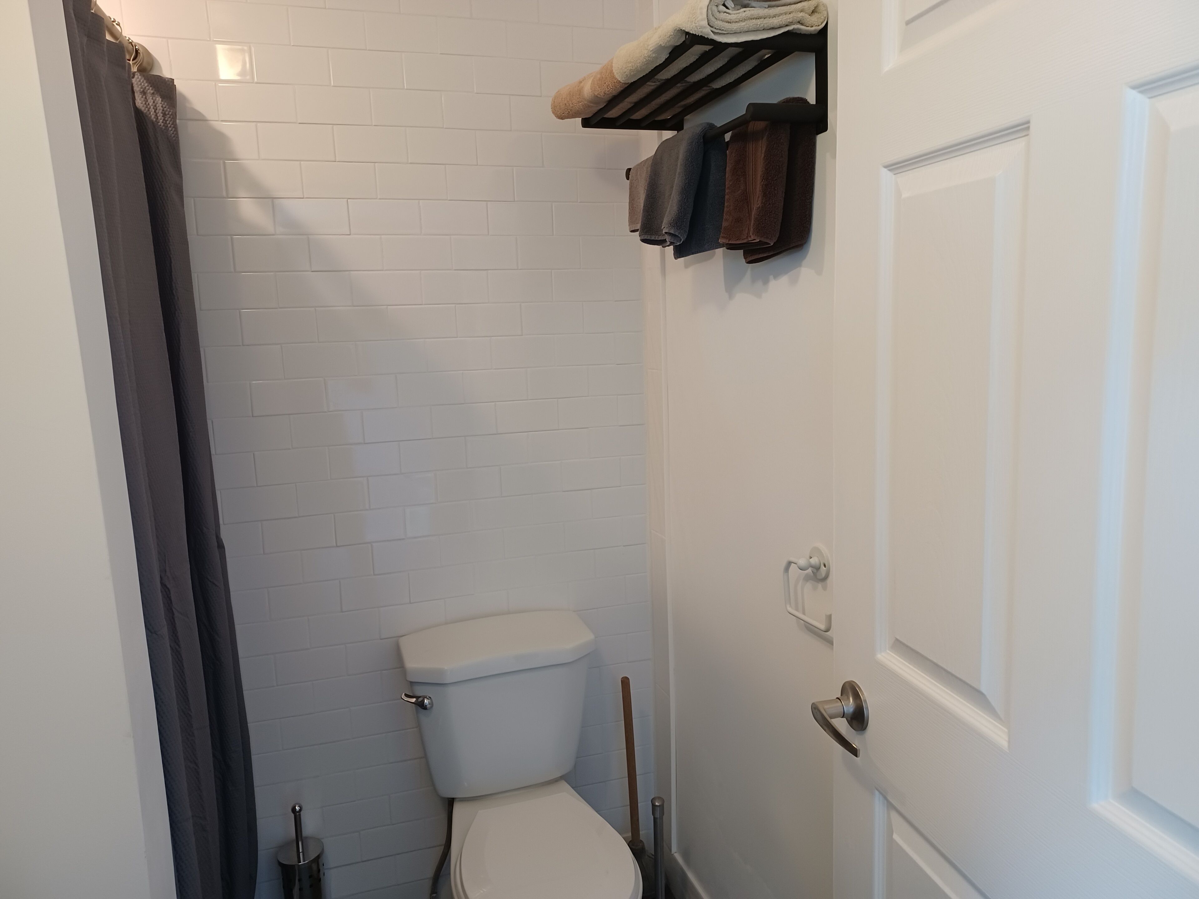 Et lille, rent badeværelse med hvide flisevægge, et hvidt toilet, en håndklæderække med håndklæder og en dør med greb.