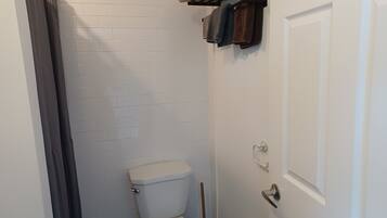 Et lille, rent badeværelse med hvide flisevægge, et hvidt toilet, en håndklæderække med håndklæder og en dør med greb.