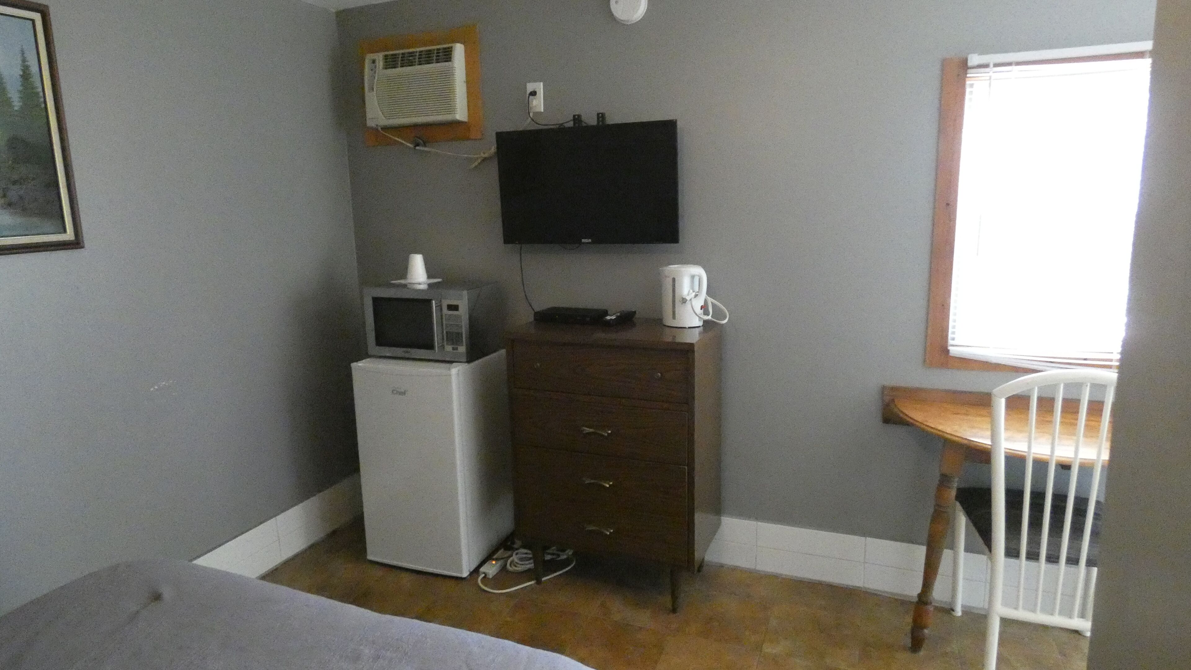 Una habitación pequeña con una cama, un escritorio con silla, un microondas, una cafetera y un televisor en la pared.