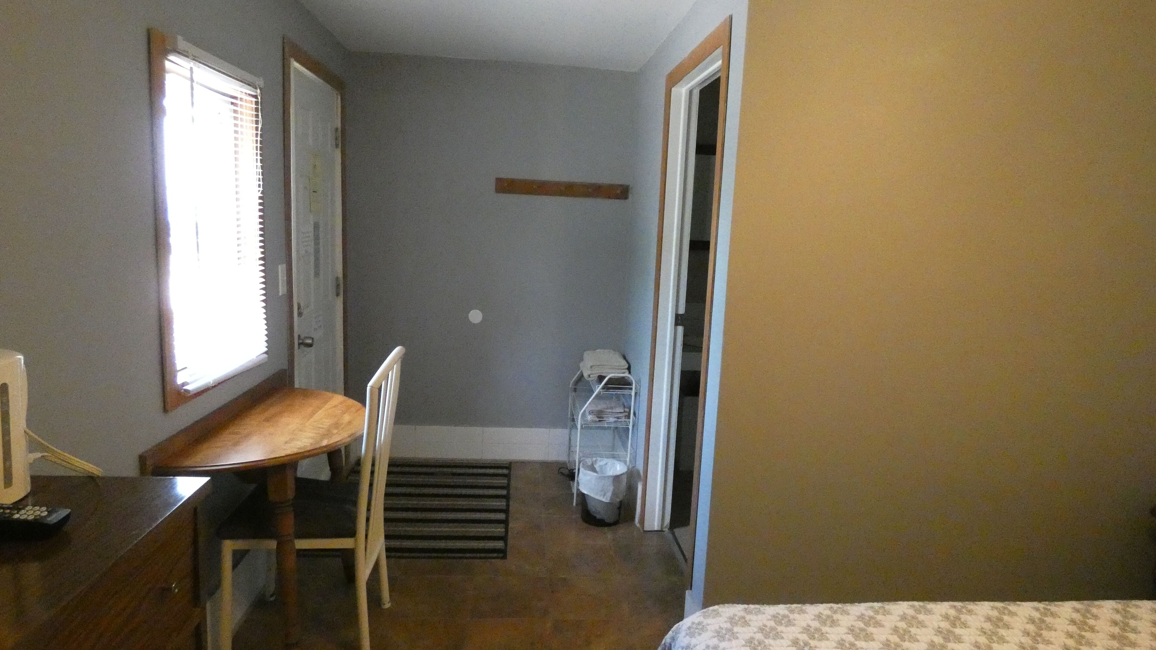 A small, compact room with a wooden desk, a chair, a radiator, a small shelf, and a door leading to another room.