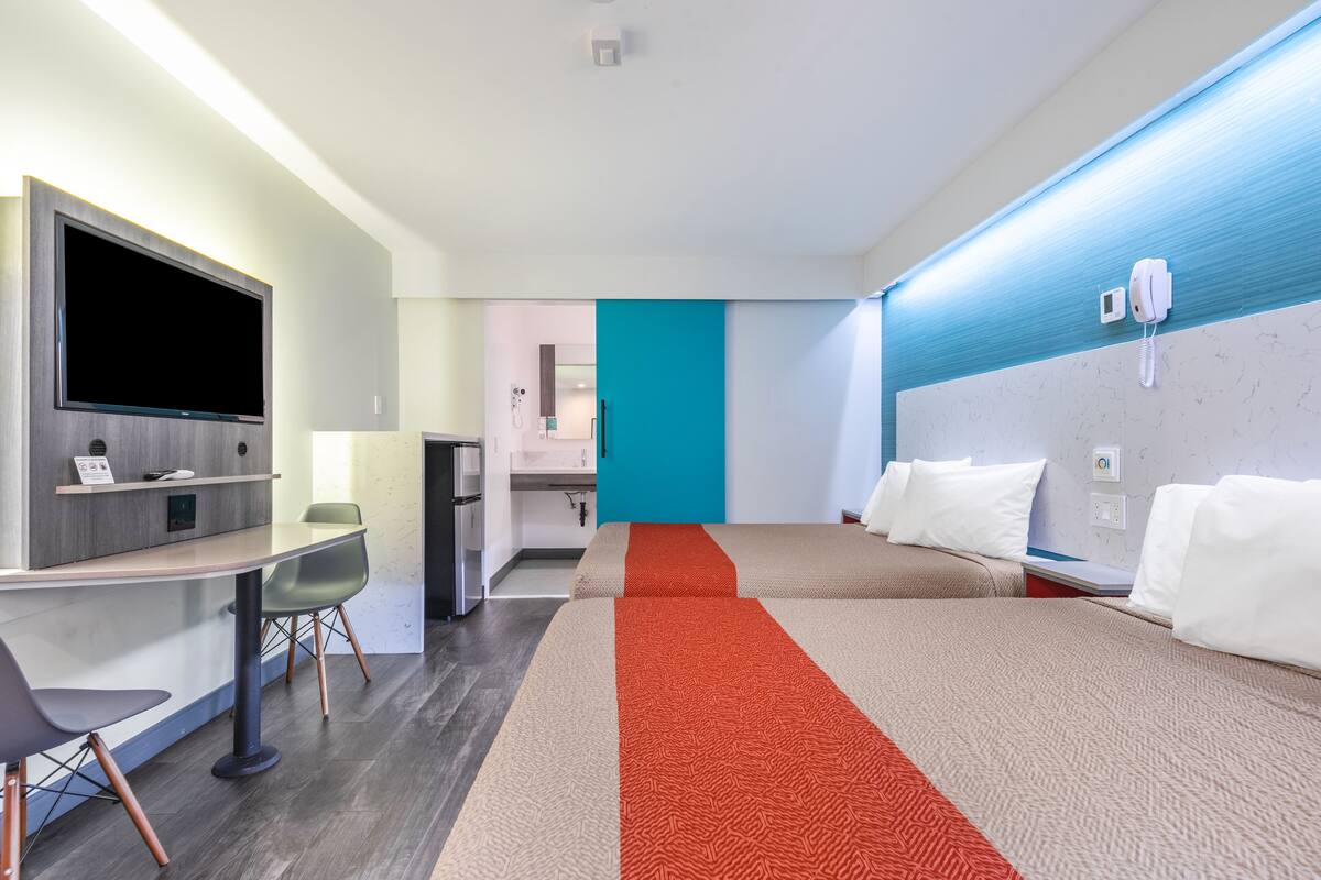 A modern hotel room with a flat-screen TV, a desk, and two beds with red and beige bedding.