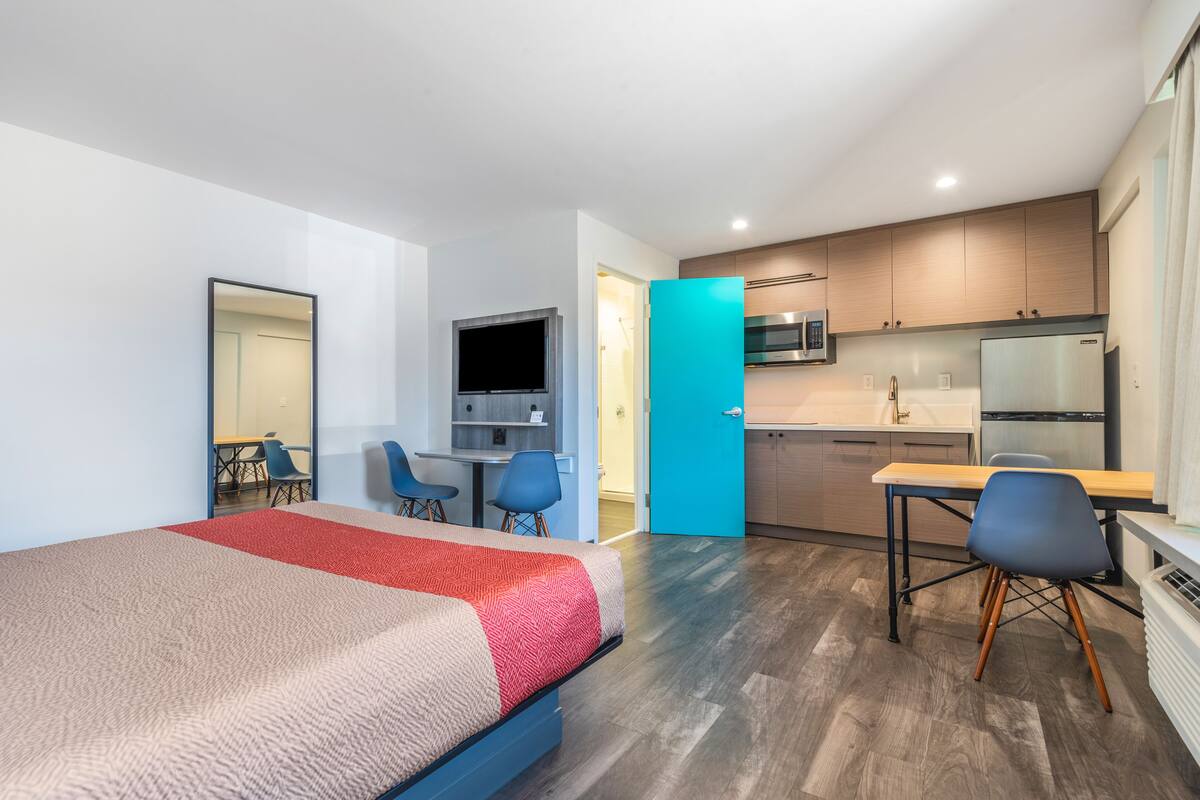 A modern hotel room with a bed, a desk, a chair, a TV, and a kitchenette.