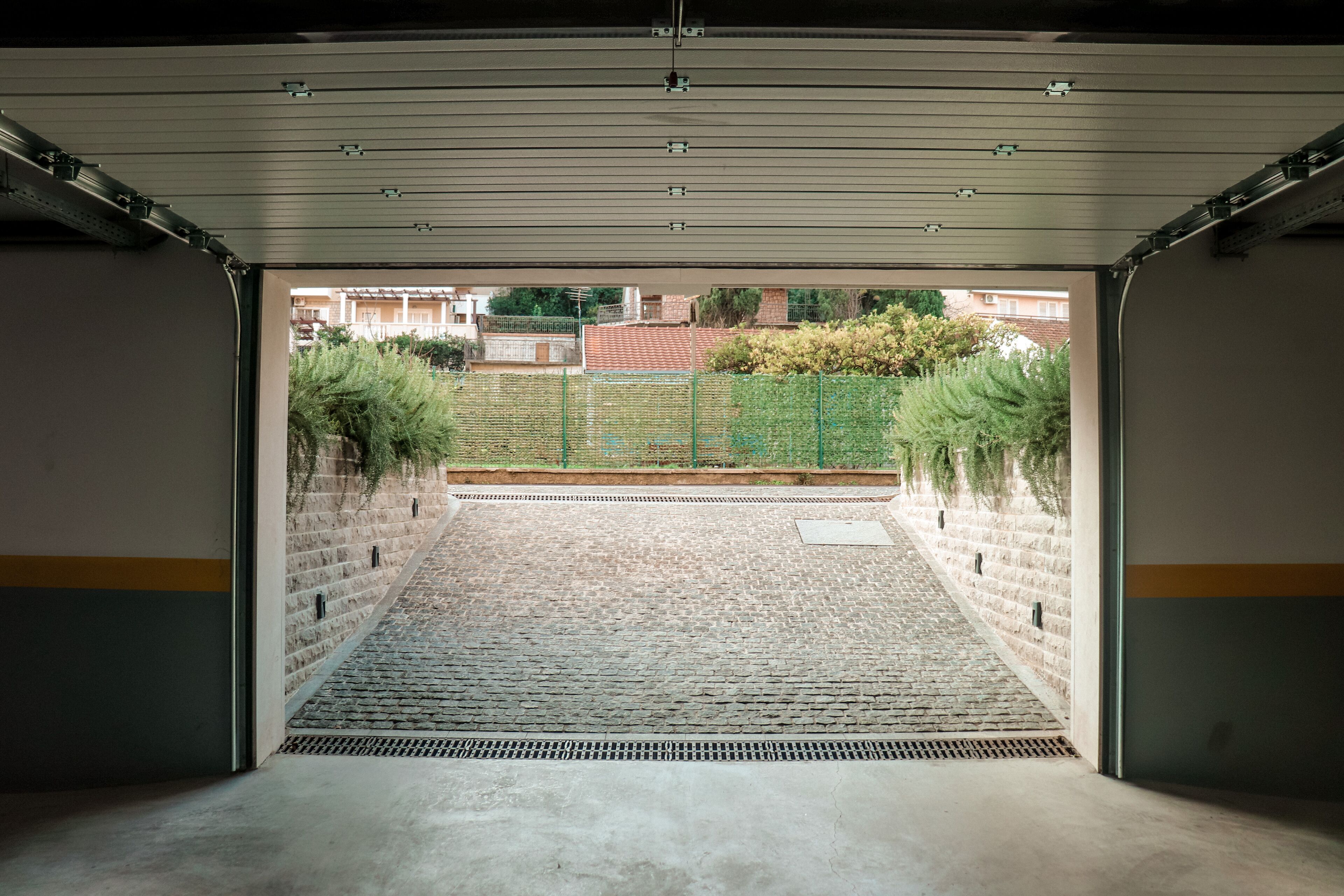 Un garage ouvert donnant sur une cour intérieure, avec un chemin en briques, une clôture verte et un mur en briques.