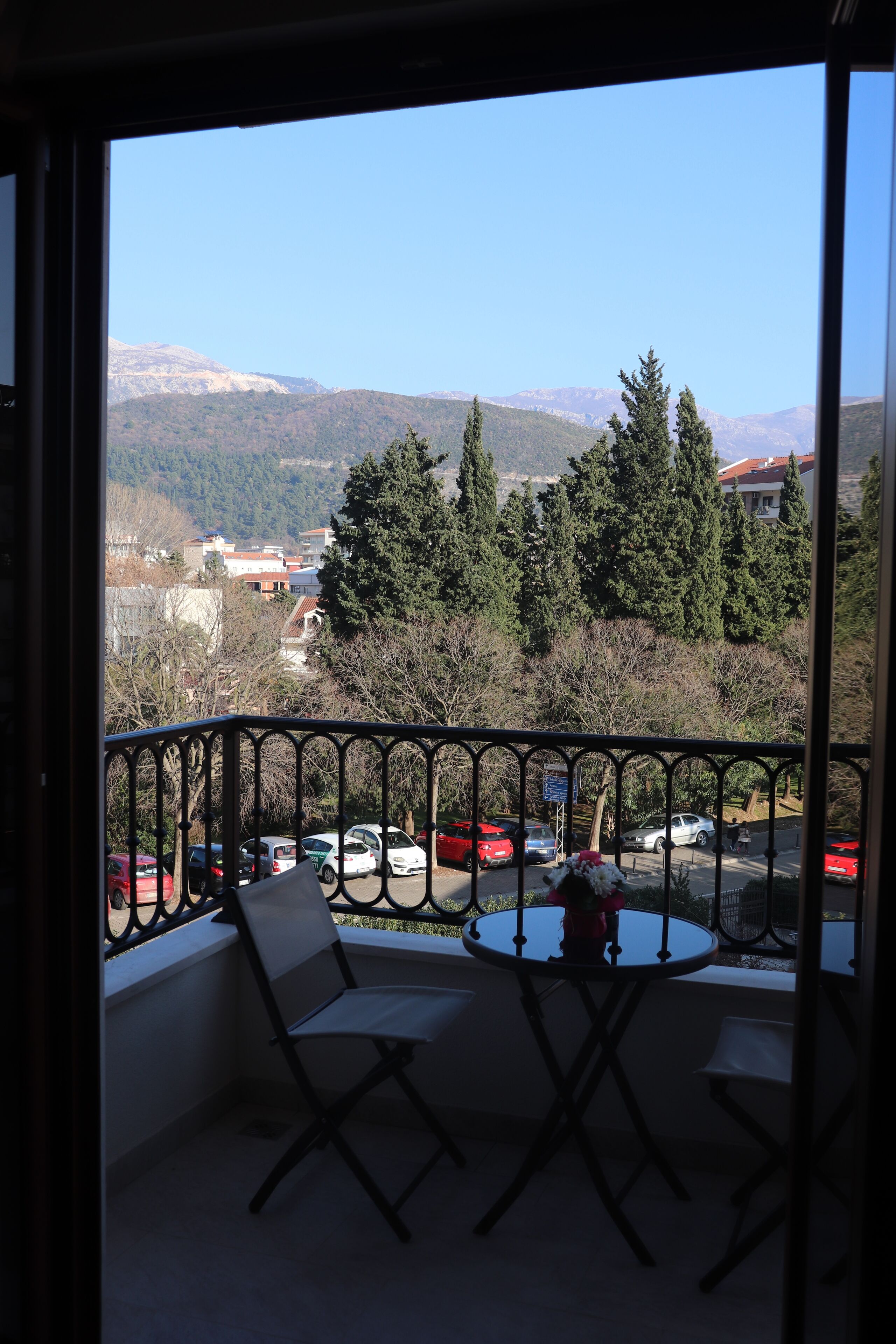 A balcony with a view of a mountainous landscape, a table with chairs, and parked cars below.