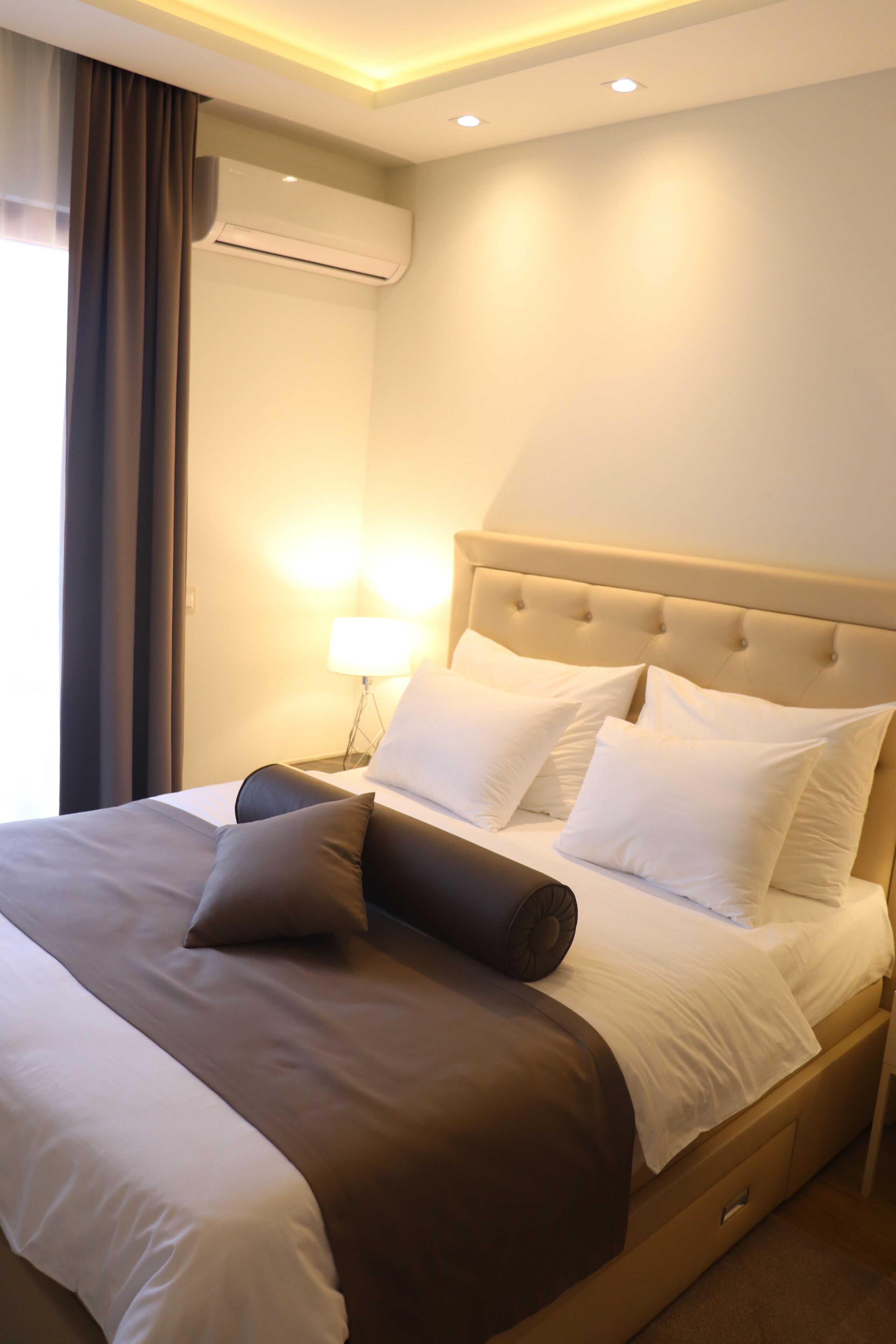 A neatly made bed with white and brown linens, a bedside lamp, and an air conditioning unit on the wall.