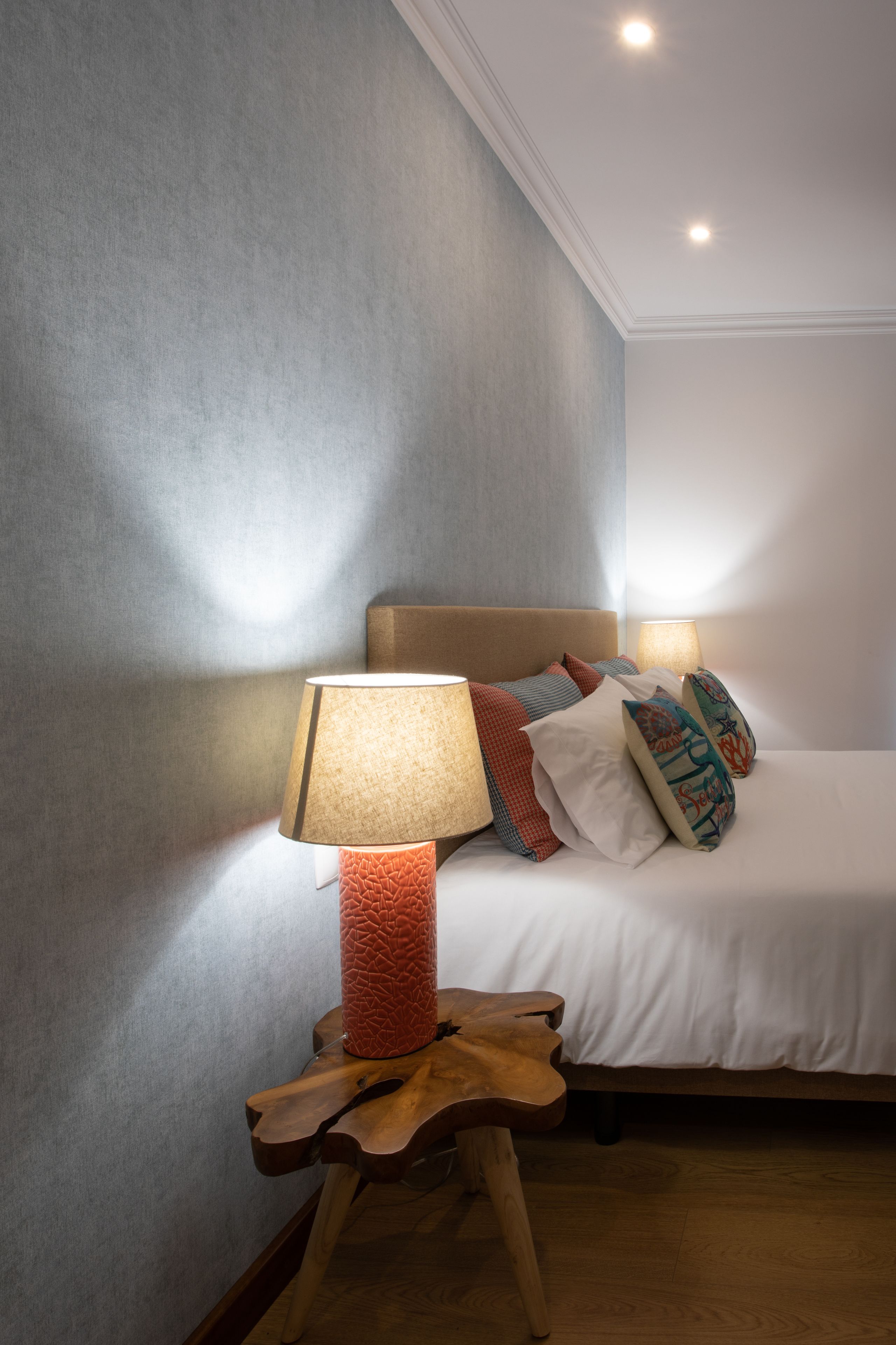 Una cama bien hecha con cabecera de madera, una lámpara roja con textura sobre un soporte de madera y una pared gris con textura.