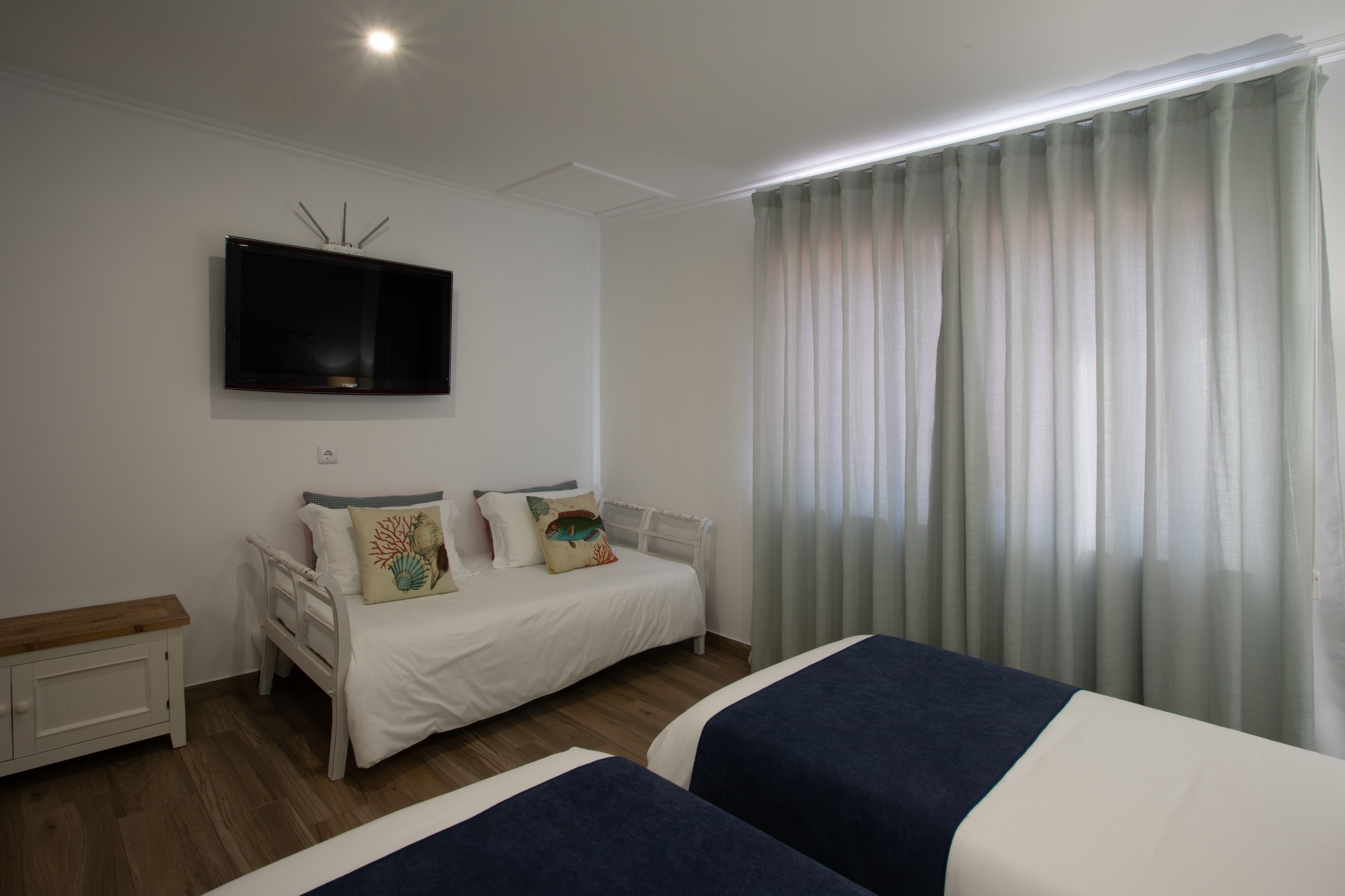 A bedroom with two beds, a flat-screen TV mounted on the wall, a bedside table, and a window with curtains.