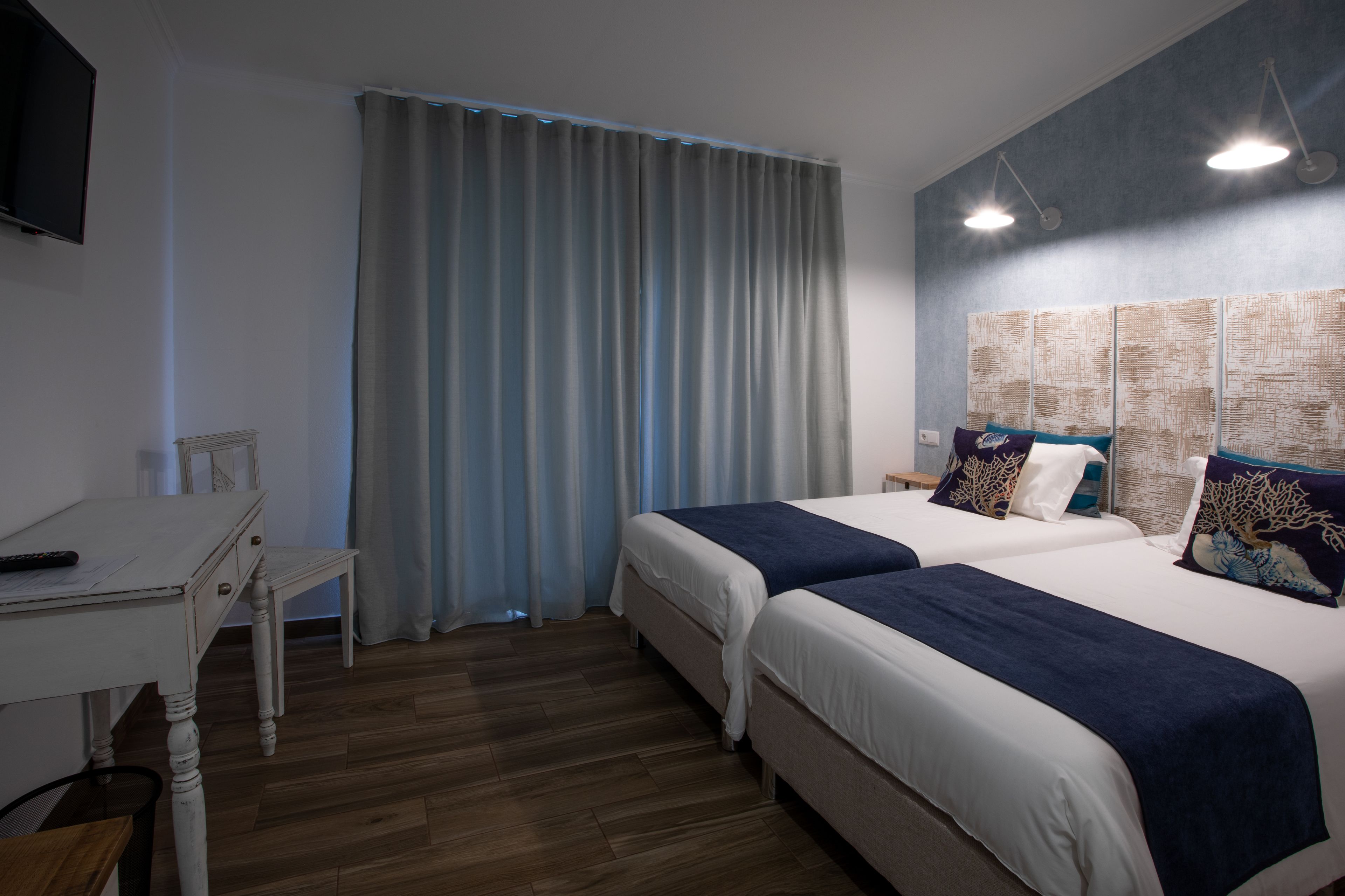 A hotel room with two beds, a desk, a TV, and a large window with curtains.