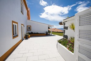 Ein modernes Haus mit weißer Fassade, einer gefliesten Terrasse und einem Garten mit verschiedenen Pflanzen.