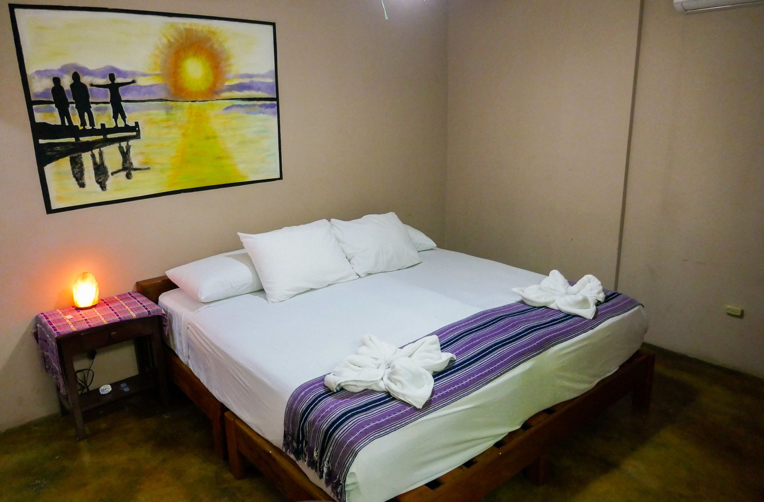 Un letto rifatto con copriletto bianco e coperta a righe viola, una lampada da comodino e un quadro raffigurante un tramonto sulla parete.