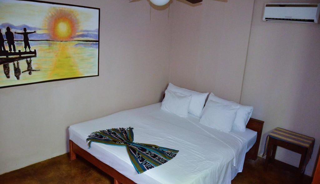 A bedroom with a bed, a painting of a sunset, an air conditioner, and a small table.