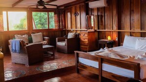 Een kamer met houten vloer, een bed, twee rieten stoelen, een houten dressoir en een plafondventilator.