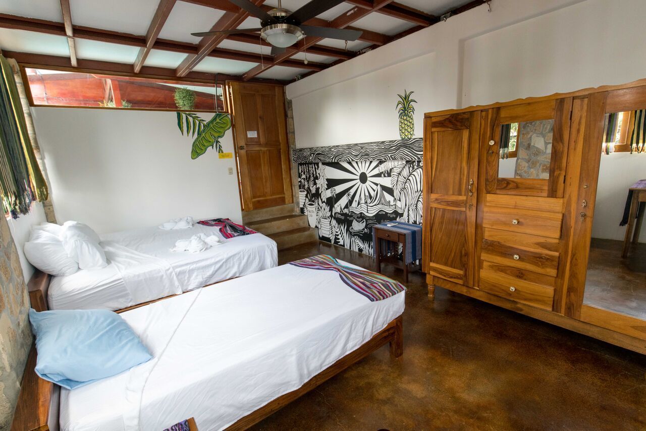 A room with two single beds, a wooden wardrobe, a mural on the wall, and a ceiling fan.