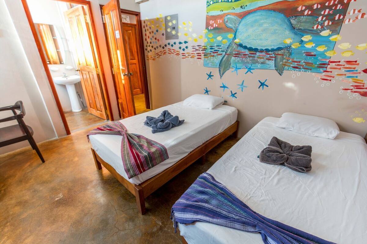 Een kamer met twee éénpersoonsbedden, een houten vloer, een badkamer met een wastafel en een muurschildering van een zeegezicht.