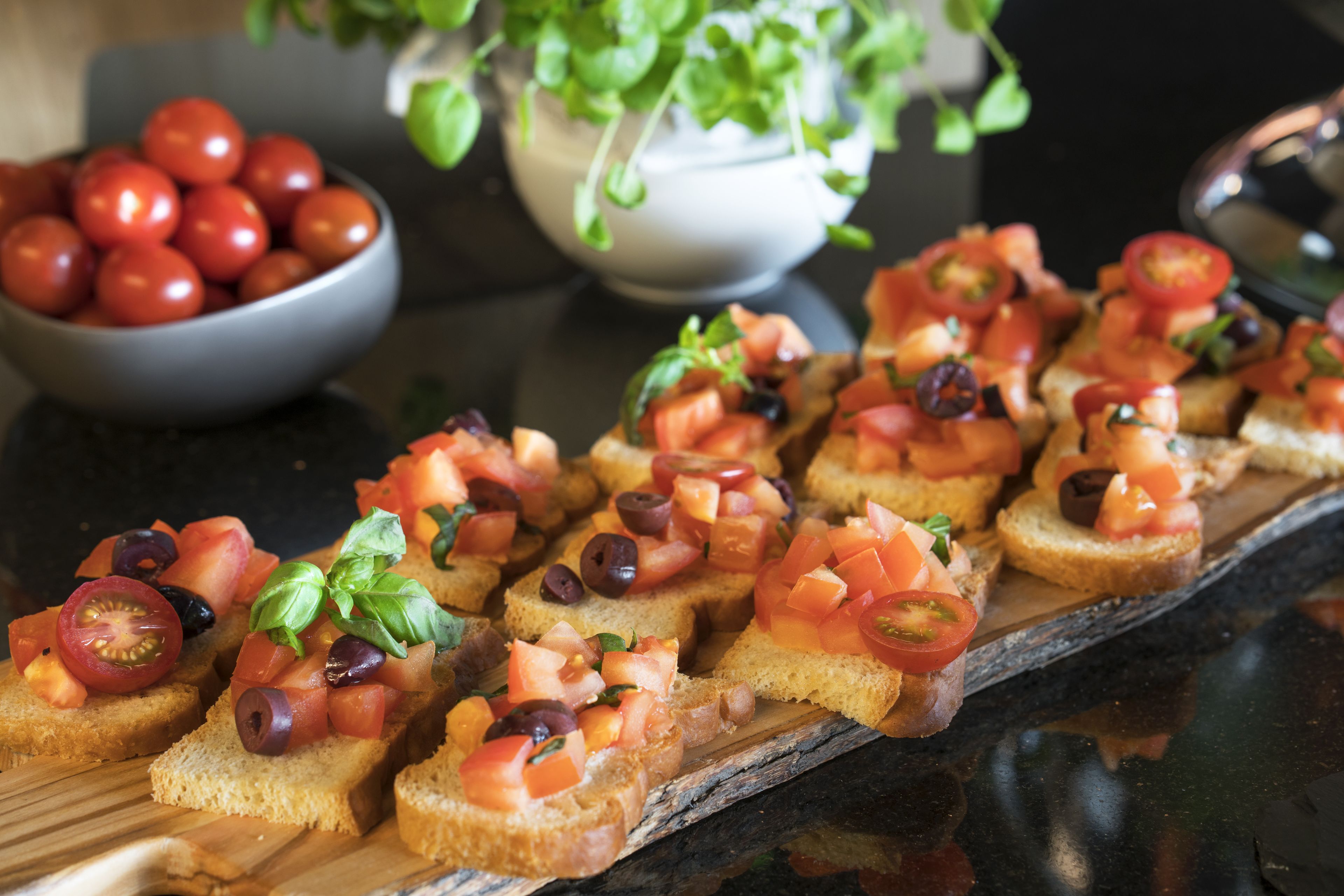 Una tabla de madera con varias tostadas cubiertas de tomates, aceitunas y albahaca.