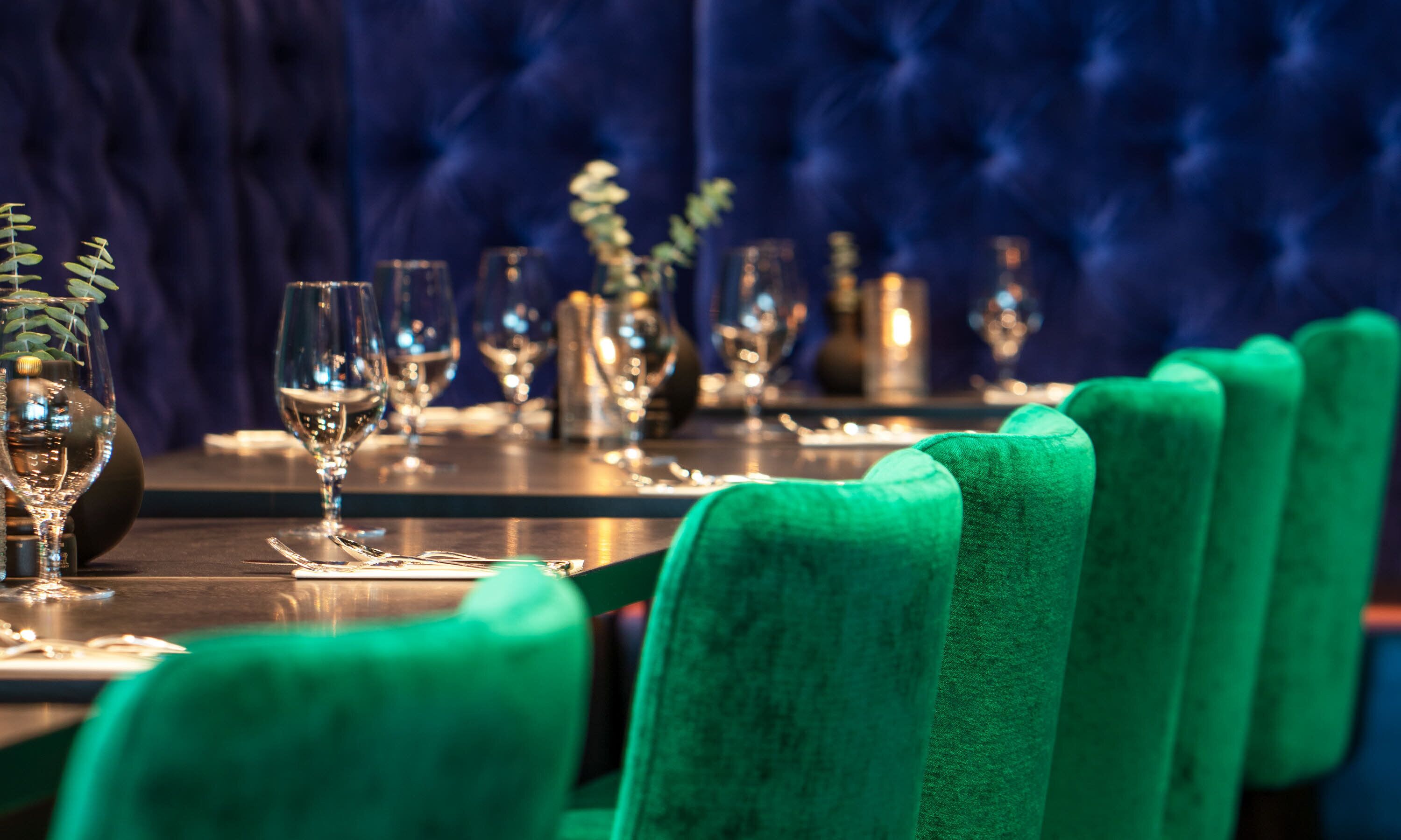 Un ambiente de restaurante con una larga mesa dispuesta para comer, sillas tapizadas en verde y varios vasos de vino.