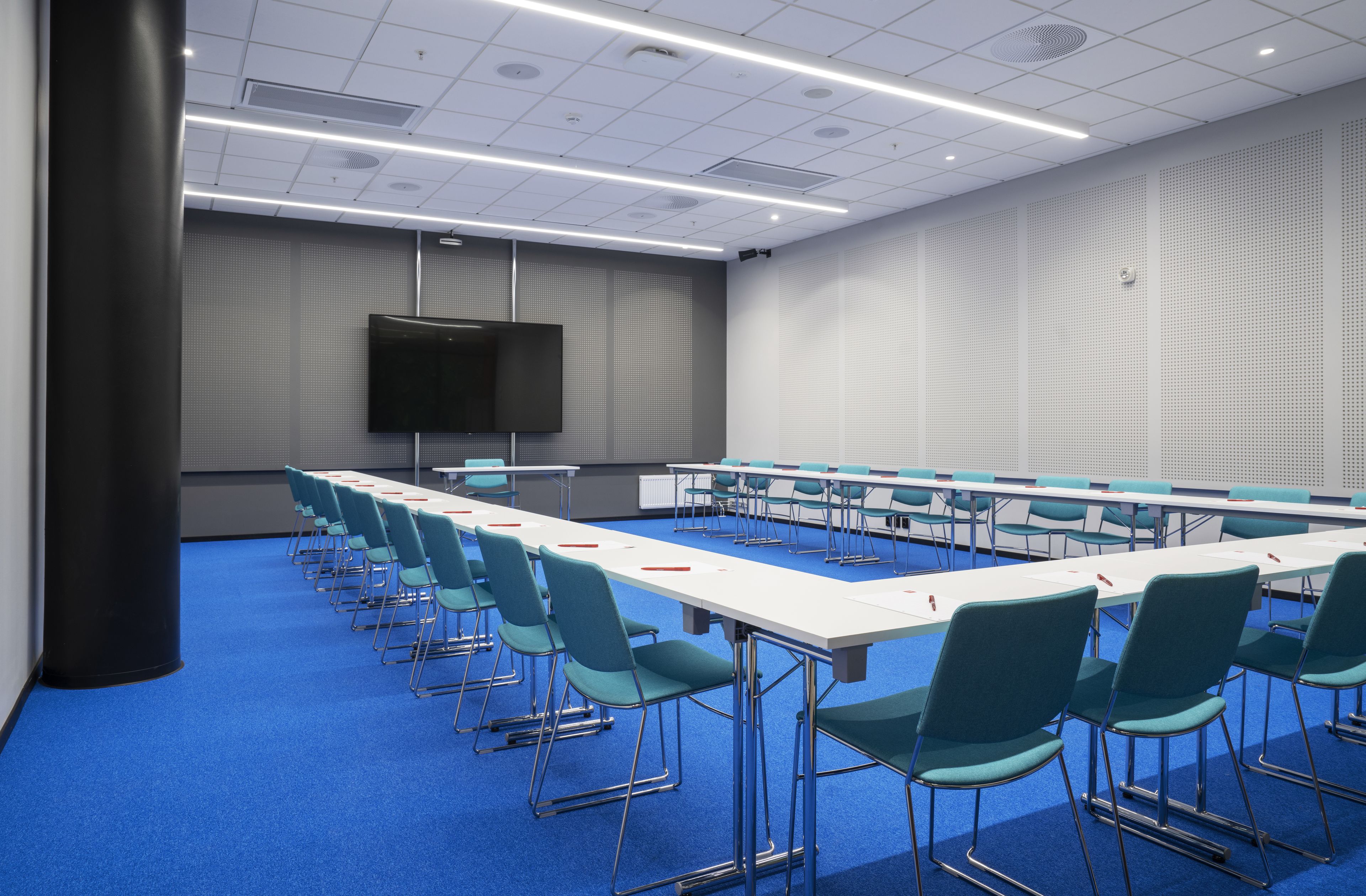 A conference room with a long table, chairs, a flat-screen TV, and a blue carpet.