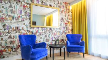 Una habitación con dos sillones azules, una mesita con un florero, un espejo grande y una pared con un mural colorido.