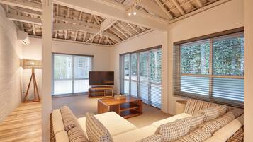 A spacious living room with a high ceiling, large windows, a sofa set, a TV, and a wooden coffee table.