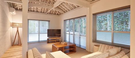 A spacious living room with a high ceiling, large windows, a sofa set, a TV, and a wooden coffee table.