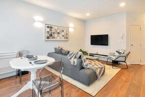 A modern living room with a grey sofa, a white dining table, and clear chairs. A flat-screen TV is mounted on the wall, and a framed collage is displayed above the sofa. - Chic & Stylish Apartment with Parking (Auckland)