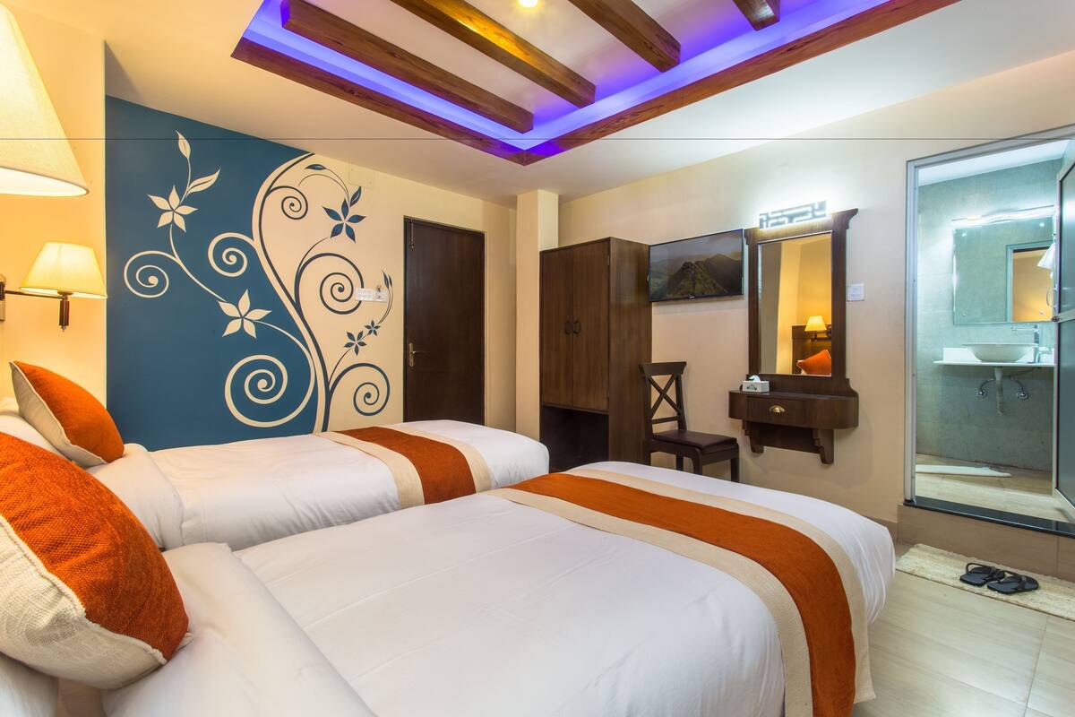 Habitación de hotel con dos camas, televisión, espejo, armario y baño visible a través de una puerta de cristal.