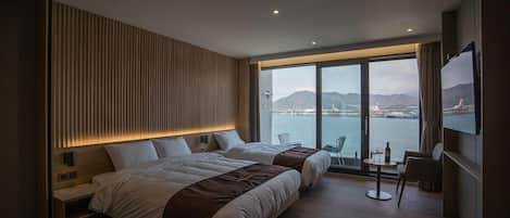 A modern hotel room with two beds, a large window with a view of the sea, a small table with a bottle and glasses, and a TV mounted on the wall.