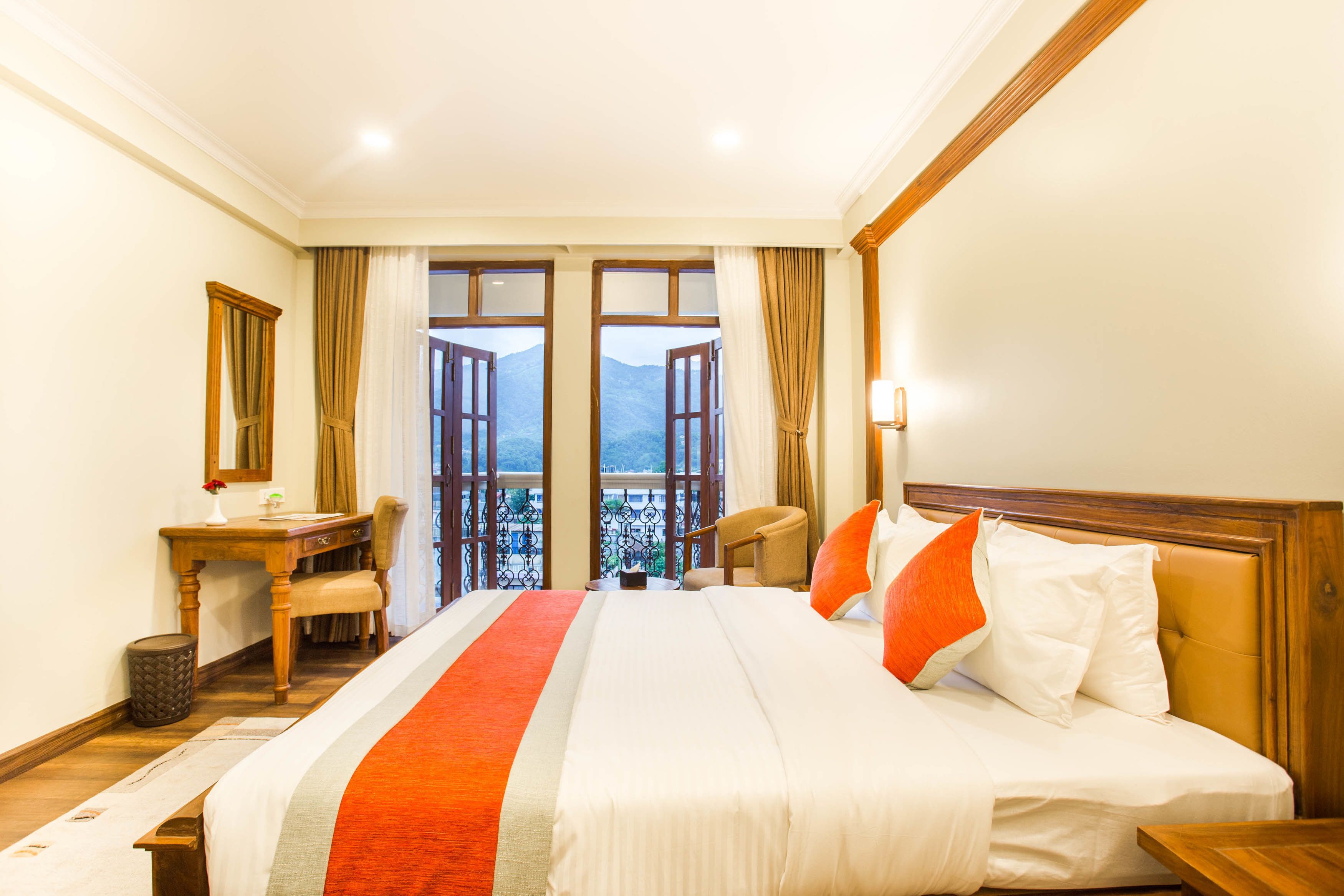 A hotel room with a large bed, a desk, a chair, and a view of mountains through open French doors.