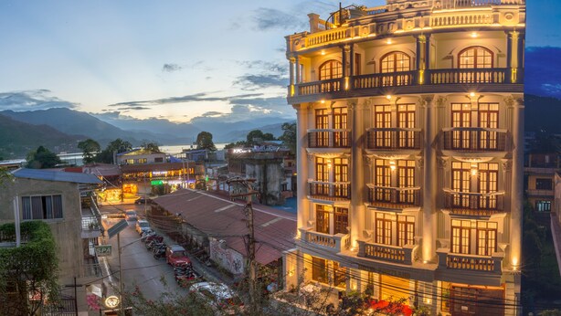 Un immeuble à plusieurs étages, doté de balcons et de grandes fenêtres, entouré d’une rue où sont garées des voitures, et offrant une vue sur des montagnes en arrière-plan.