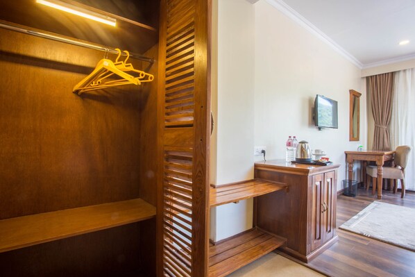 Quarto de hotel com guarda-roupa de madeira, uma pequena mesa com chaleira, uma cadeira e televisão na parede.