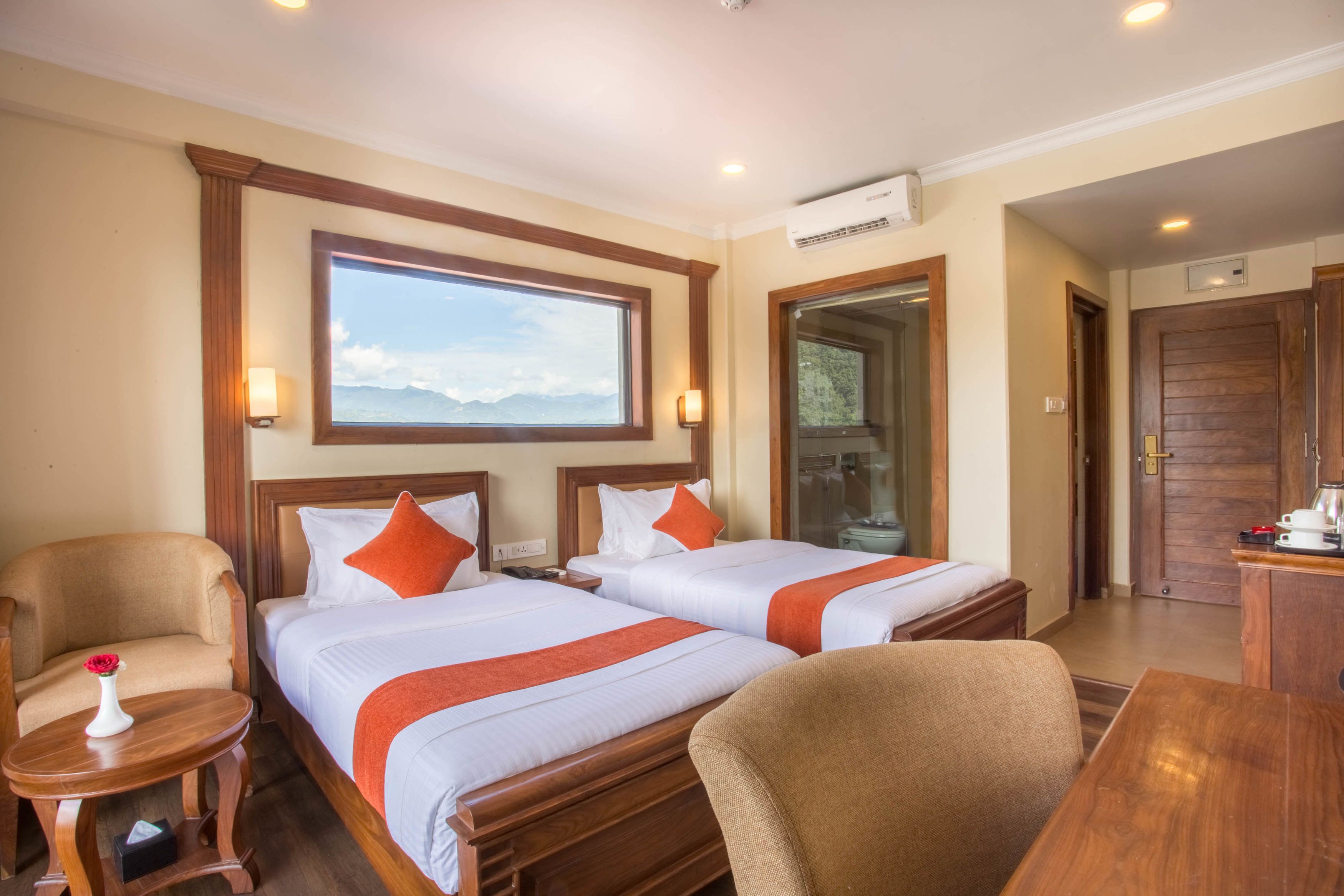 A hotel room with two beds, a wooden desk, a chair, a small table with a vase, and a window with a mountain view.