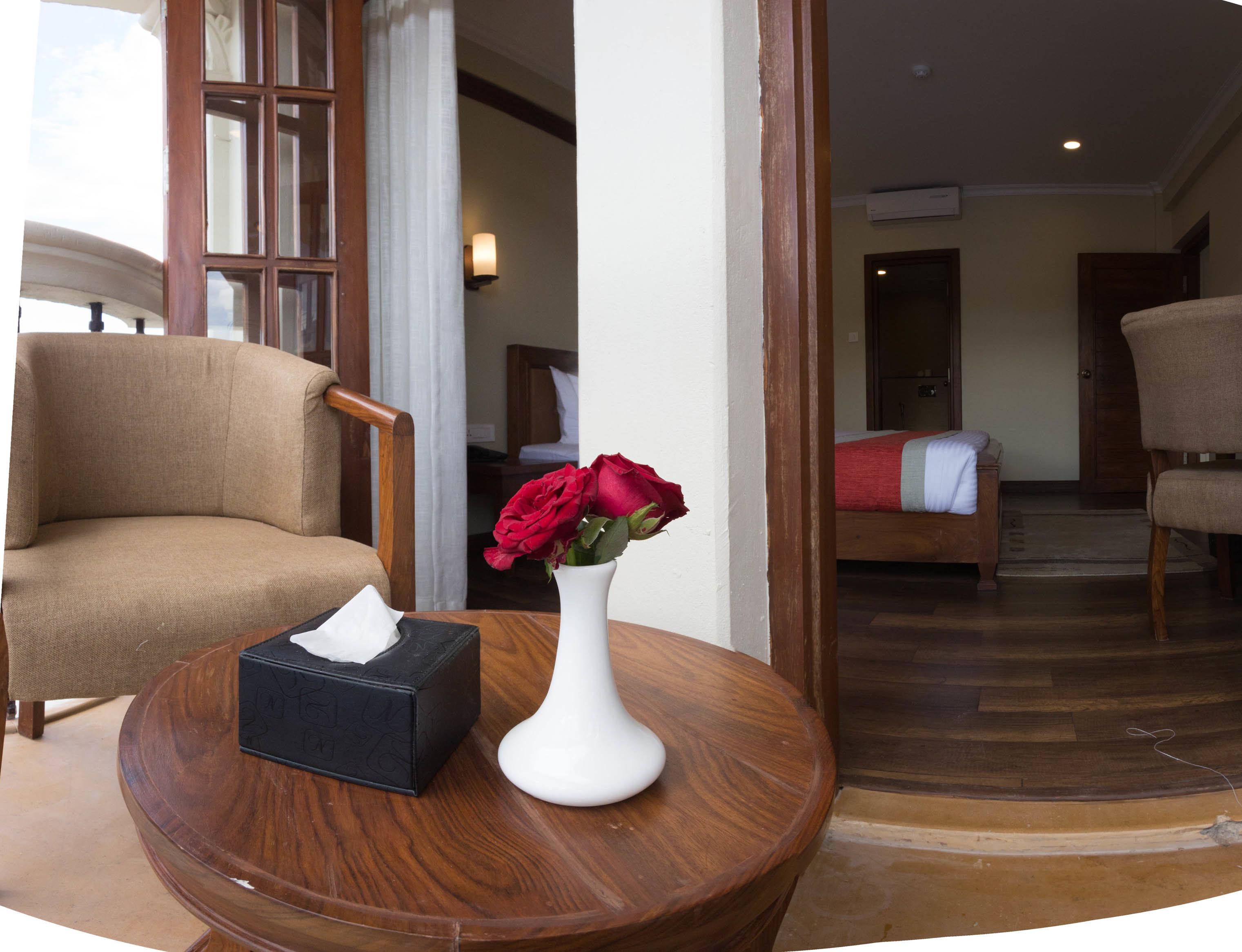 Une chambre d’hôtel avec une chaise en bois, une table de chevet sur laquelle se trouve une boîte de mouchoirs et un vase rempli de roses rouges, et un lit recouvert d’une couverture rouge.