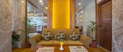 A modern living room with a sofa, patterned cushions, a wooden coffee table with newspapers, and potted plants.