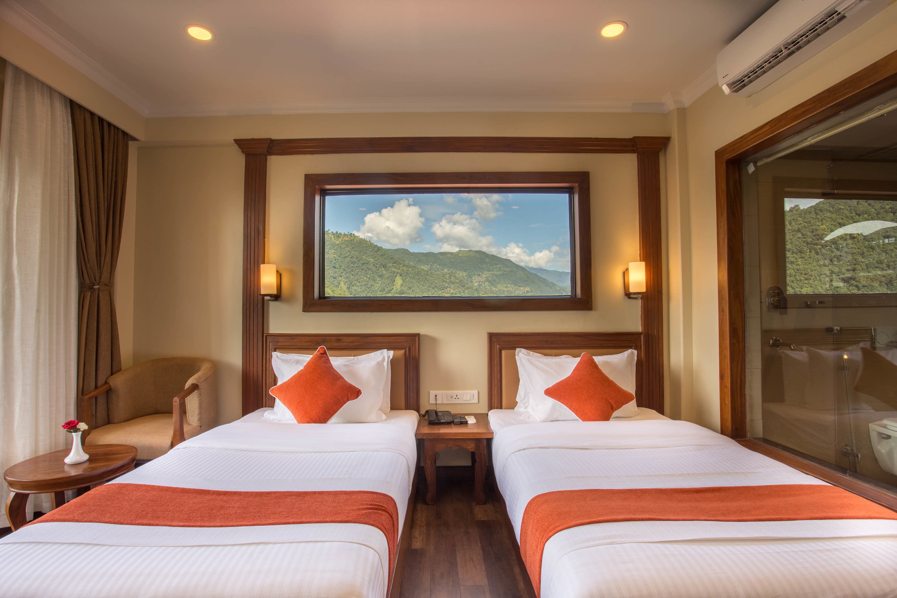 A hotel room with two beds, a nightstand, a chair, a small table with a vase, and a large window with a mountain view.