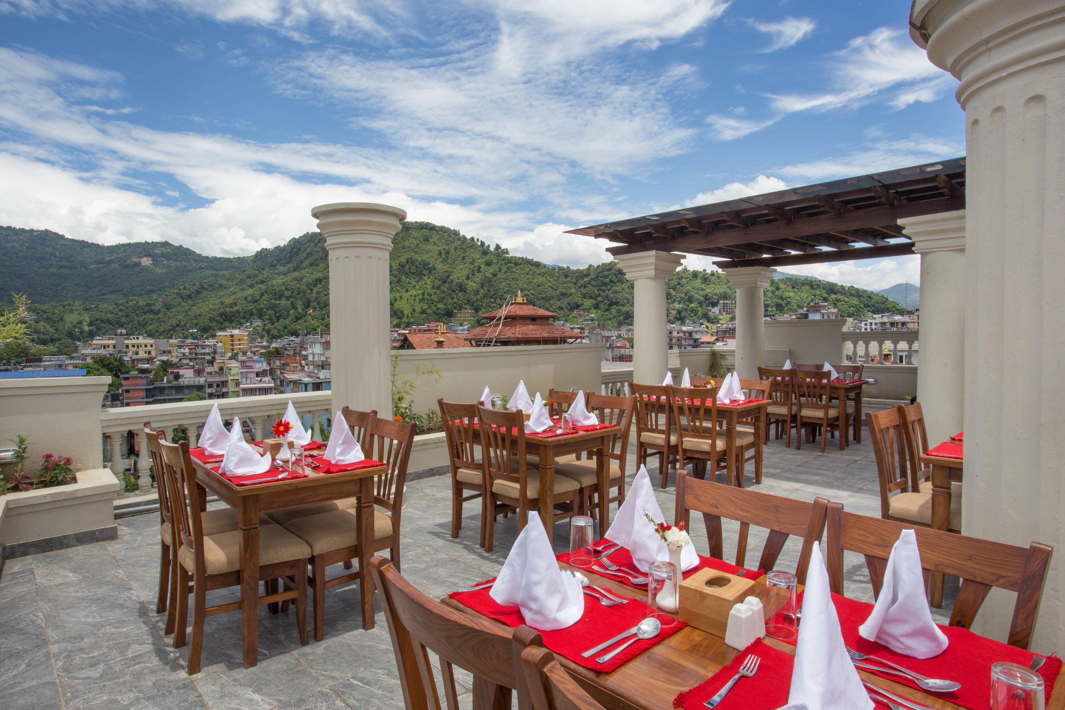 An outdoor dining area with tables set for two, wooden chairs, and a view of a cityscape and mountains in the background.