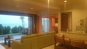 Une chambre d’hôtel moderne dotée d’une grande porte-fenêtre coulissante donnant sur un balcon, d’un canapé, d’un coin repas avec une table et des chaises, et d’une petite kitchenette.
