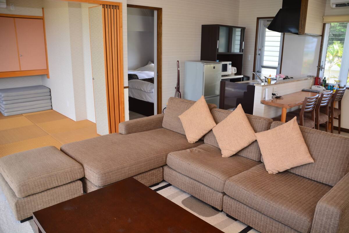 Sala de estar com sofá seccional, mesa de centro de madeira e área de cozinha compacta com micro-ondas e geladeira.