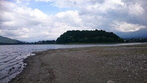 A lakeside view with a rocky shore, a small island with dense trees, and distant buildings. - Rental Villa Ooishiso (Fujikawaguchiko)