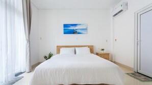 A neatly made bed with white bedding, a wooden headboard, and a small bedside table with a plant. - Linh House (Da Nang)