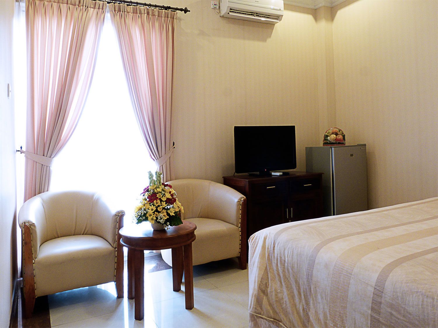 A hotel room with a bed, a TV on a wooden cabinet, a small table with a flower arrangement, two armchairs, and a refrigerator.