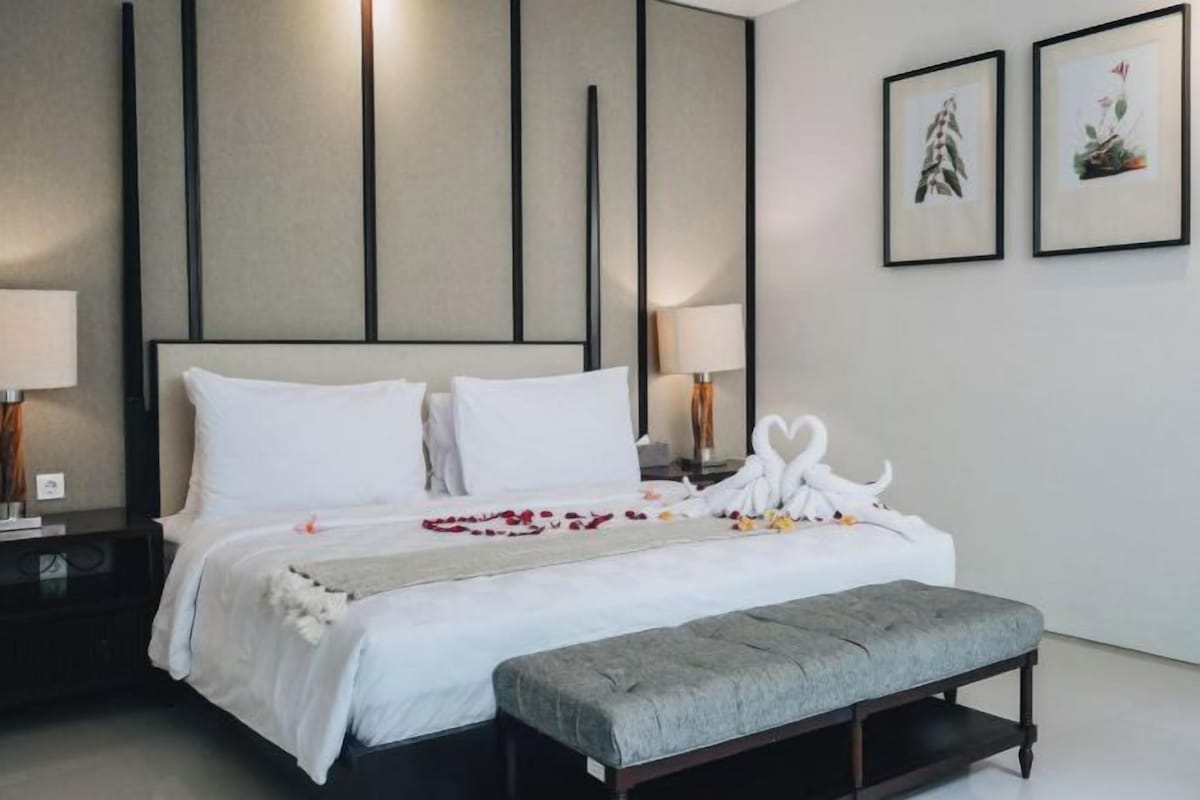 Un lit bien fait, avec du linge de lit blanc, un banc gris et deux tableaux encadrés accrochés au mur.