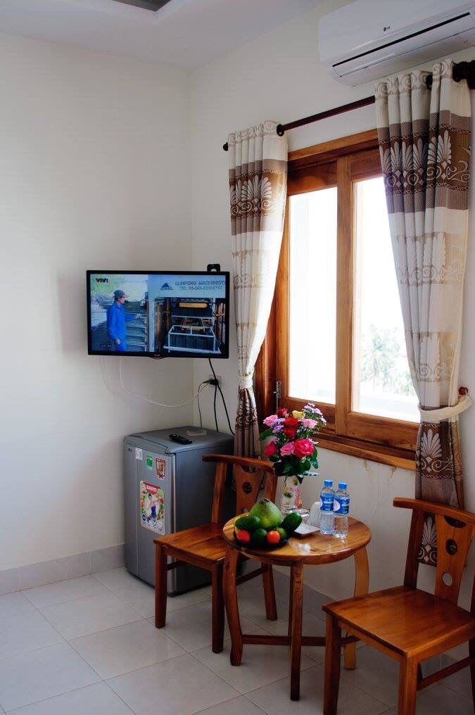 A room with a television mounted on the wall, a small refrigerator, a wooden table with chairs, and a window with curtains.