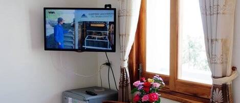 A room with a television mounted on the wall, a small refrigerator, a wooden table with chairs, and a window with curtains.