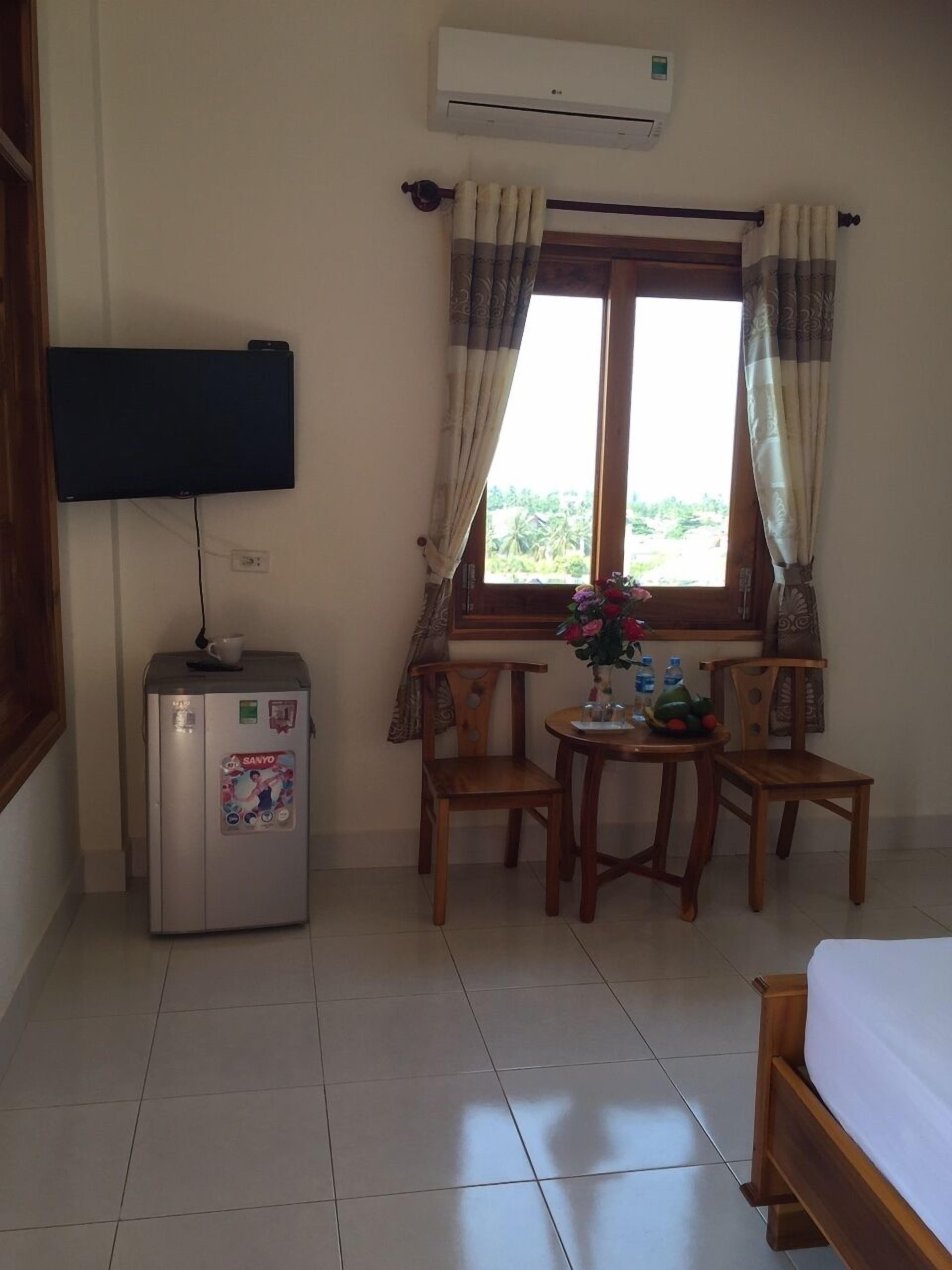 A room with a bed, a small table with chairs, a refrigerator, a window with curtains, and an air conditioning unit.