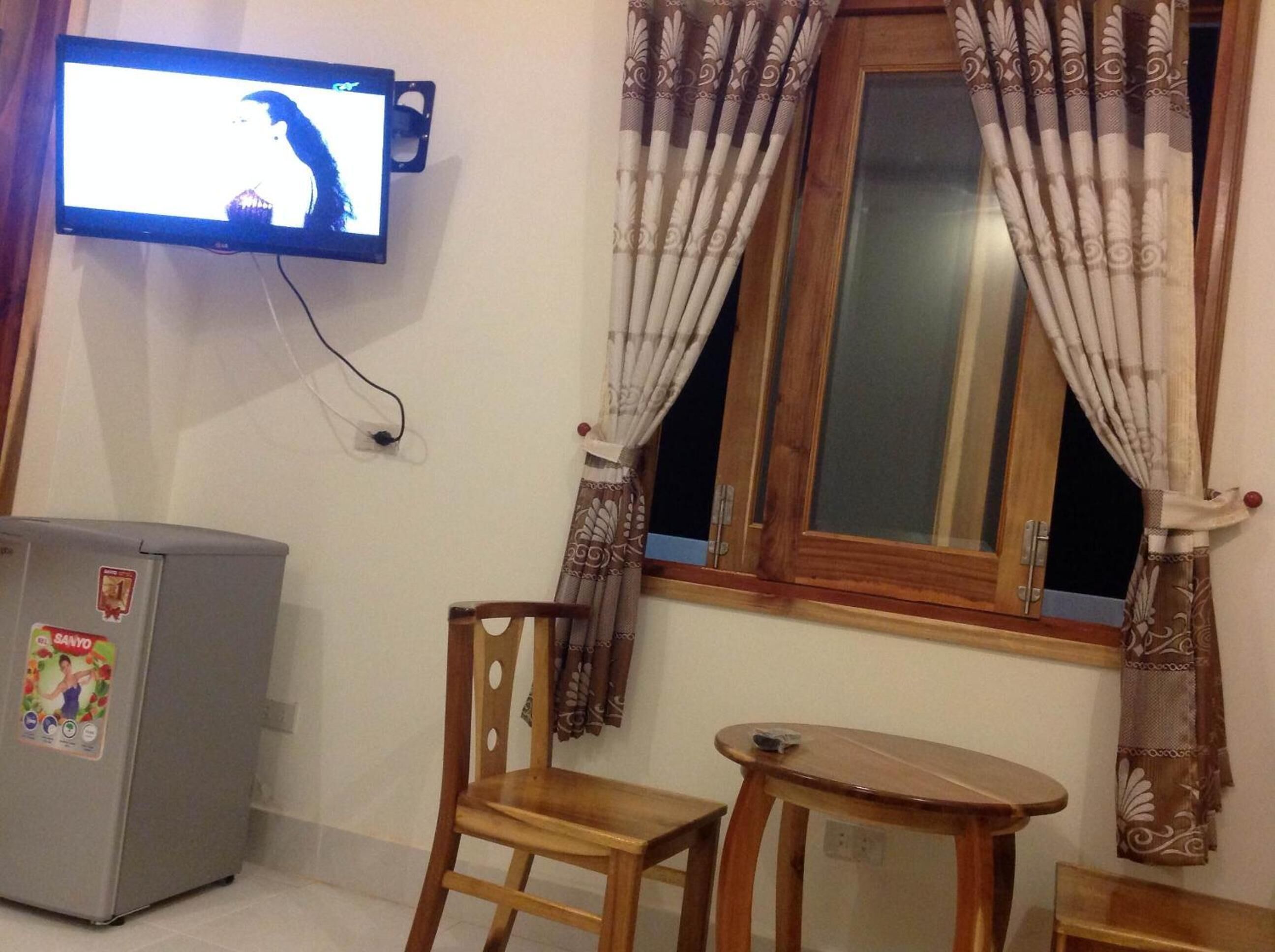 Une pièce avec un téléviseur fixé au mur, un petit réfrigérateur, une chaise en bois, une petite table et une fenêtre avec des rideaux.