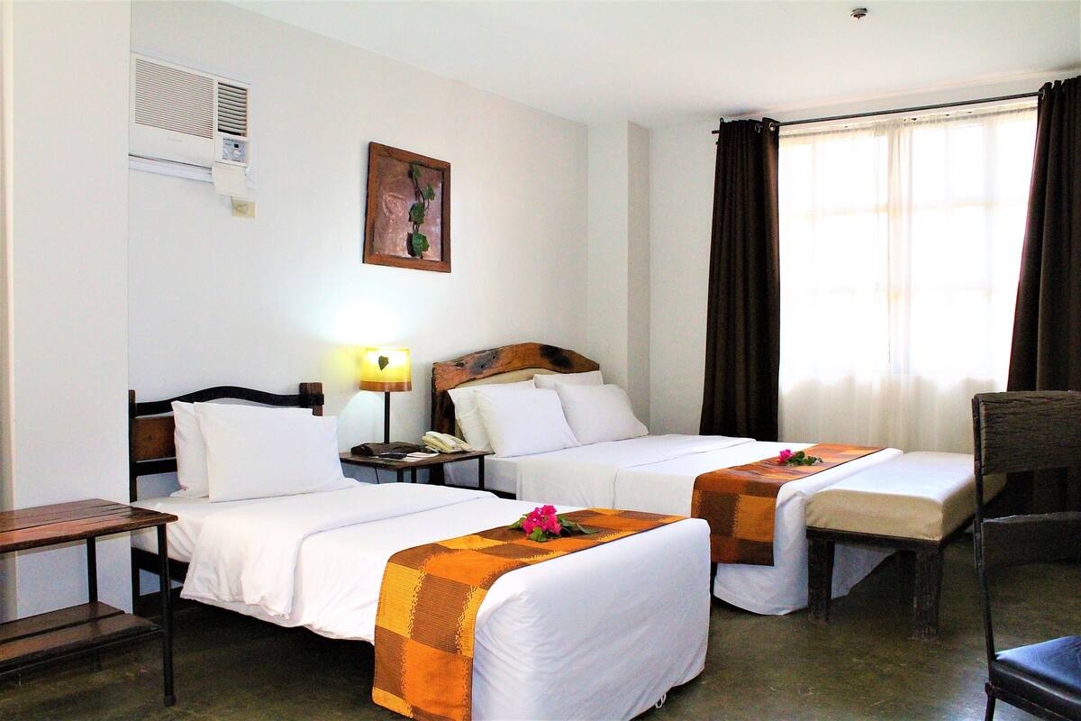 A hotel room with two beds, a wooden headboard, a nightstand with a lamp, a window with curtains, and a wooden chair.