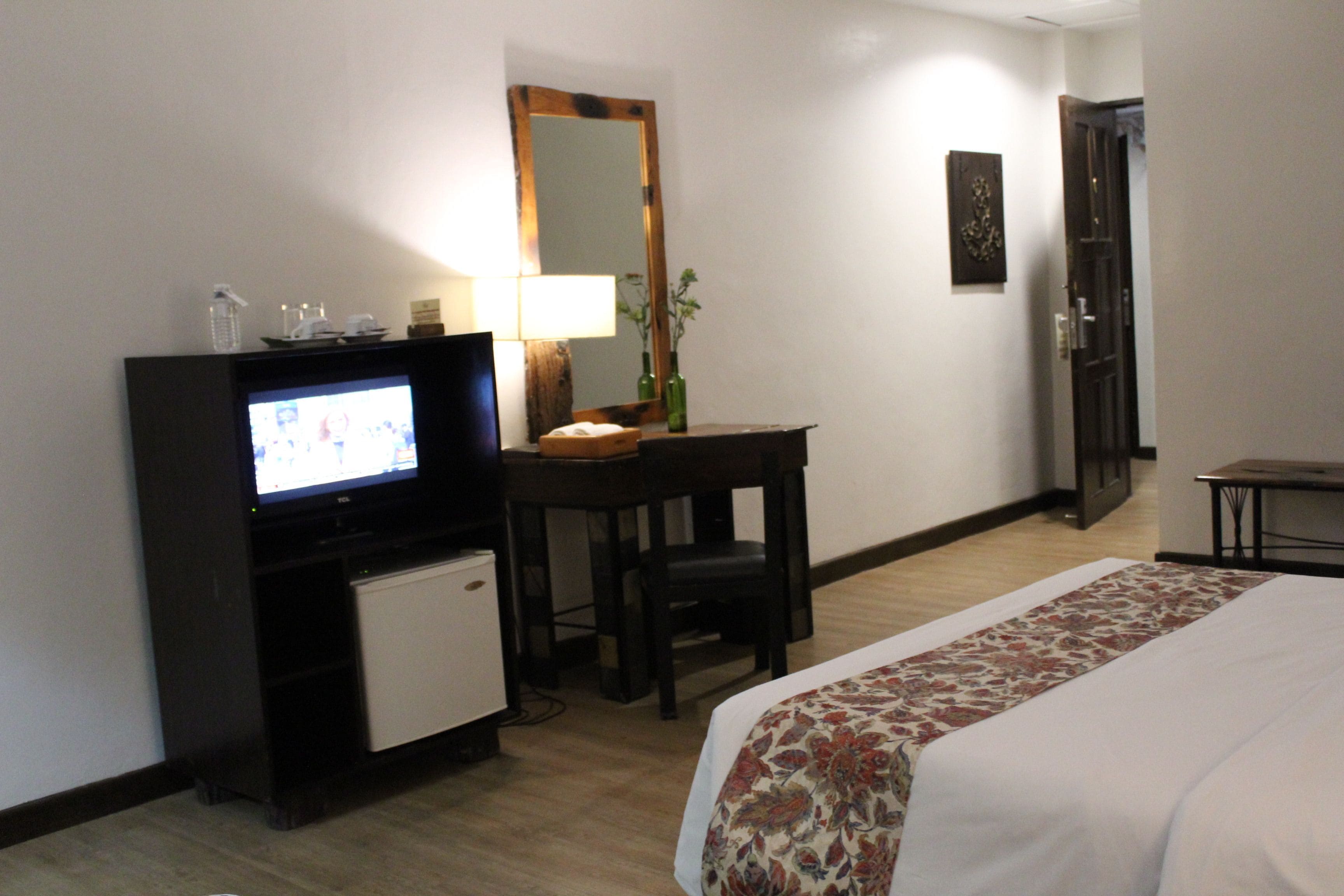 A hotel room with a bed, a desk with a television, a small refrigerator, a mirror, and a door.