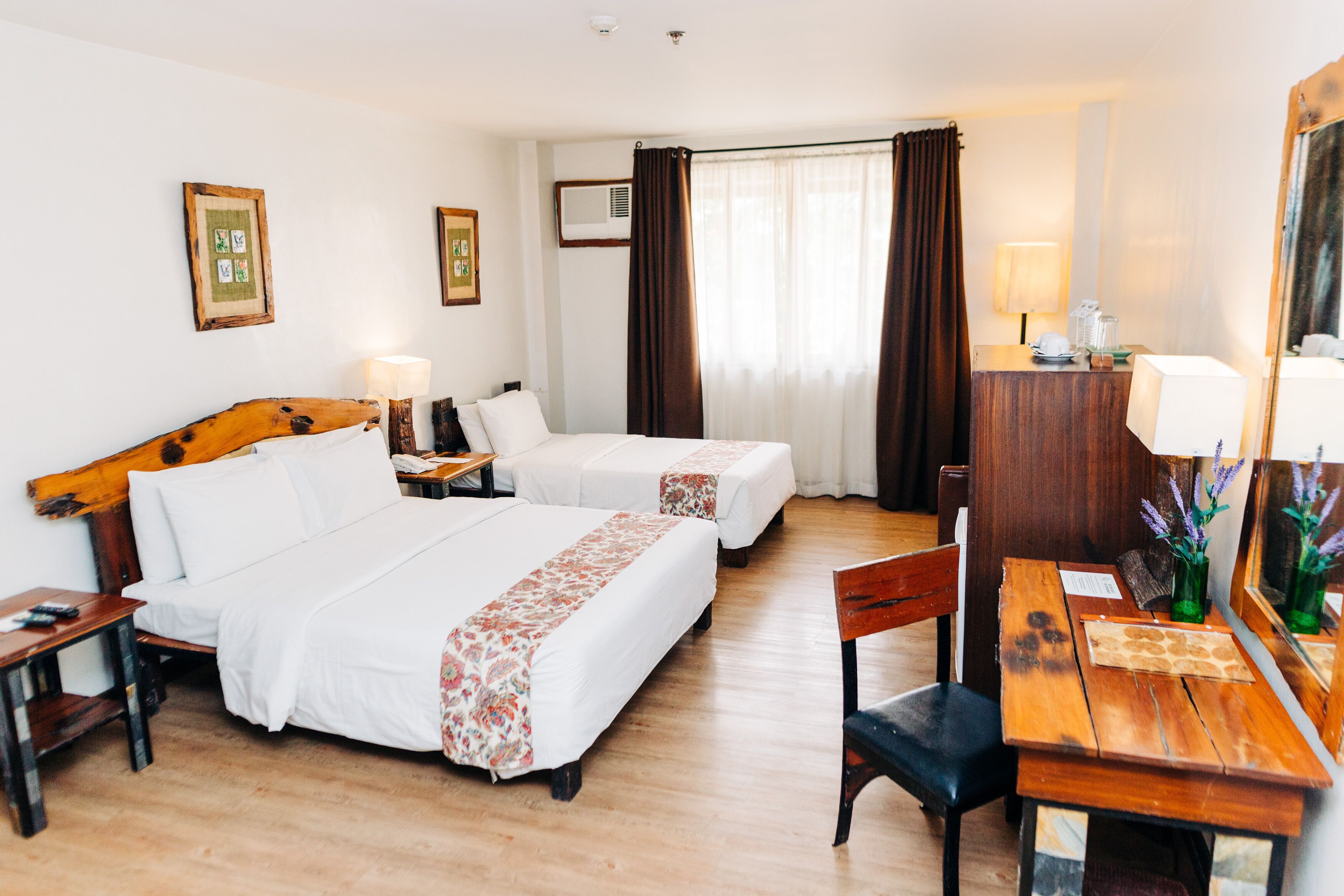 A hotel room with two beds, a wooden desk, a chair, a mirror, and a window with curtains.